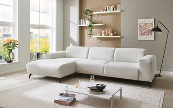 Stilvolles weißes Ecksofa in modernem Wohnzimmer mit großen Fenstern, Wandregalen und Dekoration. Rechts daneben lächelnde Frau, die ein Herz mit den Händen formt – Symbol für Wohlfühlen und Wohnkomfort.